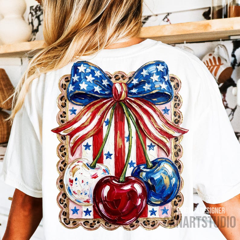May include: White t-shirt featuring a patriotic design. The design includes a blue bow with white stars, red and white striped ribbons, and three cherries. The cherries are painted in red, blue, and white with gold accents. The design is framed with a leopard print border.