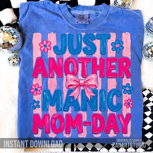 Op de afbeelding: Een blauw Comfort Colors T-shirt met de tekst "JUST ANOTHER MANIC MOM-DAY" in roze en blauw. Het ontwerp bevat roze verticale strepen, bloemen en een strik. De woorden "INSTANT DOWNLOAD" staan onderaan.