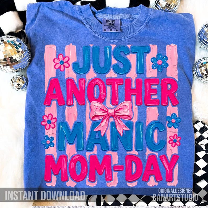 Op de afbeelding: Een blauw Comfort Colors T-shirt met de tekst "JUST ANOTHER MANIC MOM-DAY" in roze en blauw. Het ontwerp bevat roze verticale strepen, bloemen en een strik. De woorden "INSTANT DOWNLOAD" staan onderaan.
