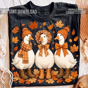 Op de afbeelding: Een donkergrijs t-shirt met drie witte ganzen tussen verspreide oranje herfstbladeren. De ganzen dragen oranje winteraccessoires, waaronder een gebreide muts, een geruite sjaal, een bloemenkapje en bruine laarzen. Tekst: INSTANT DOWNLOAD.