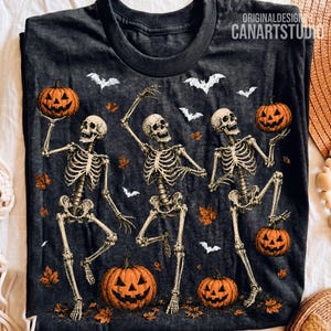 May include: A charcoal gray t-shirt featuring a graphic print of three skeletons dancing among orange jack-o'-lanterns and white bats. The design includes scattered orange autumn leaves and the text ORIGINALDESIGN CANARTSTUDIO at the top.