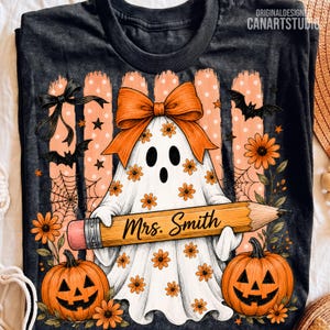 Puede incluir: Una camiseta gris oscuro con una ilustraci&oacute;n de un fantasma blanco con un lazo naranja, sosteniendo un l&aacute;piz amarillo grande con la etiqueta "Mrs. Smith". El dise&ntilde;o incluye calabazas naranjas, murci&eacute;lagos negros, telara&ntilde;as y flores.