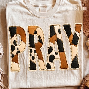 Könnte beinhalten: Ein cremefarbenes T-Shirt mit dem Wort PRAY in großen Blockbuchstaben. Jeder Buchstabe ist mit einem Patchwork aus braunen, schwarzen und weißen Kuhfellmustern gefüllt und mit einem Rand aus kleinen, runden, weißen Perlen verziert.