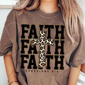 May include: A brown t-shirt with the word "FAITH" repeated three times in black with a leopard print cross in the center. The text "EPHESIANS 2:8" is printed below the word "FAITH".