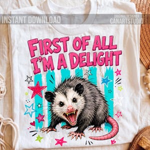 Op de afbeelding: Een wit t-shirt met een afbeelding van een grommende opossum en de tekst "FIRST OF ALL I'M A DELIGHT" in roze letters. Het ontwerp bevat verticale blauwgroene strepen, diverse kleurrijke sterren in roze, blauw en groen, en kleine hartjes.