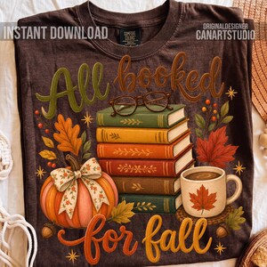 以下が含まれることがあります： ダークブラウンのTシャツで、5冊の本の積み重ねの上に眼鏡、花柄の蝶結びがついたカボチャ、カエデの葉が描かれたマグカップのグラフィックがプリントされています。テキストは「All booked for fall」と書かれています。デザインには秋の葉と星が含まれています。