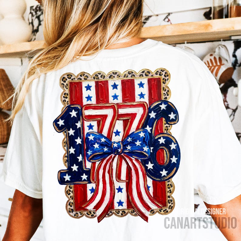 May include: White t-shirt featuring a patriotic design with the number "1776" in red, white, and blue. The design includes stars, stripes, and a bow, all within a leopard print border. The shirt is a comfortable fit.