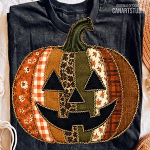 Peut inclure: Un t-shirt gris foncé avec une grande applique centrale en forme de citrouille faite de divers morceaux de tissu. La citrouille présente un visage en tissu noir avec des yeux triangulaires et une bouche dentelée, entourée de motifs floraux.