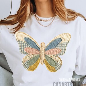May include: White t-shirt featuring a colorful butterfly applique. The butterfly design incorporates various fabric patterns and colors, including blue gingham, floral, and solid pastel hues. The text "ORIGINAL DESIGNER CANARTS STUDIO" is printed at the bottom.