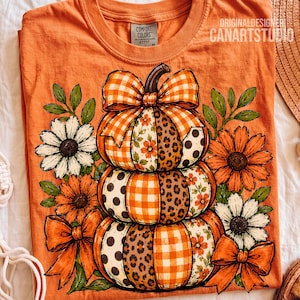 Puede incluir: Una camiseta naranja con un gráfico de tres calabazas apiladas decoradas con varios patrones, incluyendo cuadros vichy naranjas, estampado de leopardo, lunares y motivos florales. El diseño incluye lazos naranjas y flores blancas y naranjas.