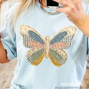 May include: Light blue t-shirt featuring a patchwork butterfly design. The butterfly has wings in various patterns and colors, including gingham, floral, and solid fabrics. The text "ORIGINAL DESIGNER CANARTSTUDIO" is visible at the bottom.