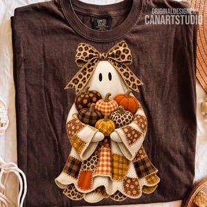 May include: A dark brown t-shirt featuring a graphic of a ghost wearing a leopard print bow and holding a cluster of fabric pumpkins. The ghost is dressed in a patchwork-style robe with various autumn-themed patterns including plaid and floral prints.
