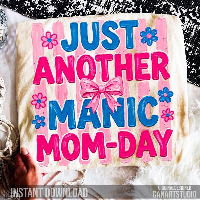 Op de afbeelding: Een cr&egrave;mekleurig T-shirt met de tekst "JUST ANOTHER MANIC MOM-DAY" in blauw en roze, versierd met bloemen en een strik. De woorden staan tegen roze verticale strepen. De woorden "INSTANT DOWNLOAD" staan onderaan.