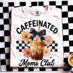 May include: Cream-colored t-shirt with the words "Caffeinated Moms Club" in black script. The design includes a graphic of an iced coffee, flowers, and a black bow. The shirt is on a black and white checkered background.