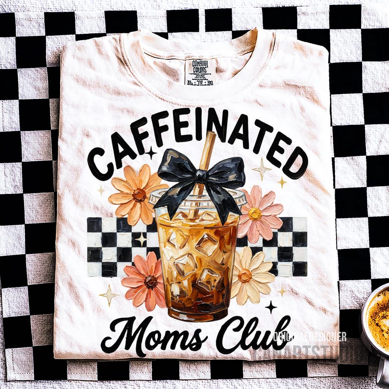 May include: Cream-colored t-shirt with the words "Caffeinated Moms Club" in black script. The design includes a graphic of an iced coffee, flowers, and a black bow. The shirt is on a black and white checkered background.