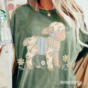 Puede incluir: Camiseta verde oliva con un dise&ntilde;o de cordero de patchwork. El cordero est&aacute; hecho de telas florales, a rayas y a cuadros. Flores bordadas rodean al cordero. El texto "ORIGINAL DESIGNER CANARTSTUDIO" est&aacute; impreso en la parte inferior derecha.