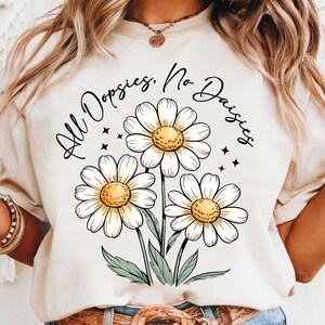 May include: Cream-colored t-shirt featuring a floral design of three daisies with white petals and yellow centers. The text "All Dopsies, No Daisies" is arched above the flowers in black script. Green leaves and black star accents complete the design.