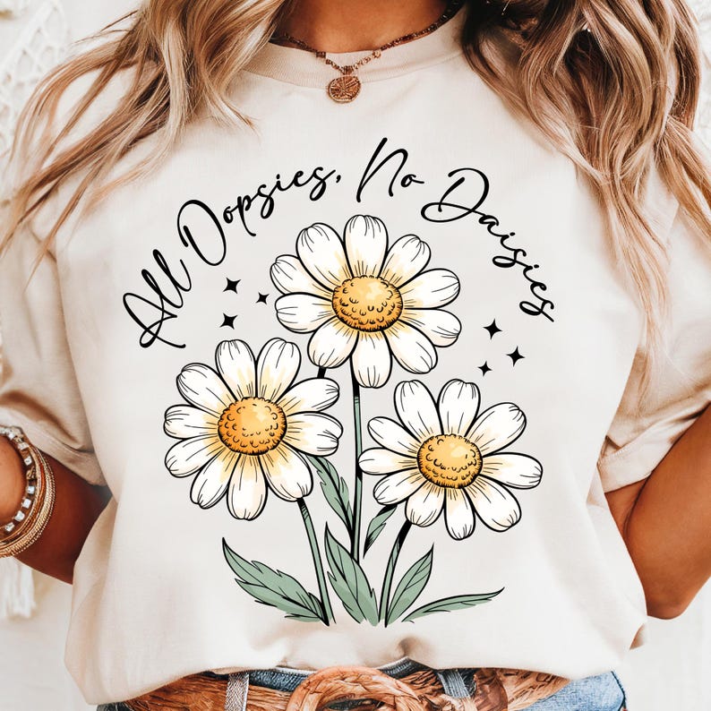 May include: Cream-colored t-shirt featuring a floral design of three daisies with white petals and yellow centers. The text "All Dopsies, No Daisies" is arched above the flowers in black script. Green leaves and black star accents complete the design.