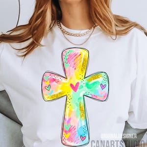 Op de afbeelding: Wit T-shirt met een kleurrijk kruisontwerp. Het kruis is versierd met roze, gele, blauwe en groene penseelstreken, gouden glitteraccenten en kleine hartjes. De tekst "CANARTS STUDIO" is zichtbaar aan de onderkant.