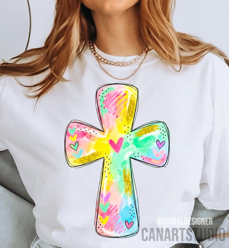 Op de afbeelding: Wit T-shirt met een kleurrijk kruisontwerp. Het kruis is versierd met roze, gele, blauwe en groene penseelstreken, gouden glitteraccenten en kleine hartjes. De tekst "CANARTS STUDIO" is zichtbaar aan de onderkant.