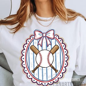 May include: White t-shirt featuring a baseball design. The design includes a baseball with red stitching, crossed wooden bats, and a bow. The design is framed by a red, white, and blue oval with a scalloped edge. The text "CANARTS STUDIO" is visible.