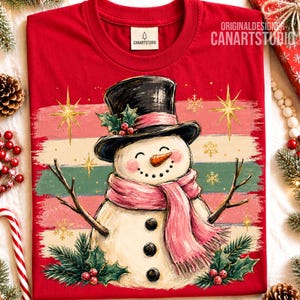 Może przedstawiać: Czerwony t-shirt z grafiką bałwana w czarnym cylindrze i r&oacute;żowym szaliku. Bałwan ma nos z marchewki, gałązkowe ramiona i trzy czarne guziki. Tło zawiera poziome paski, złote gwiazdki oraz gałązki ostrokrzewu z czerwonymi owocami.