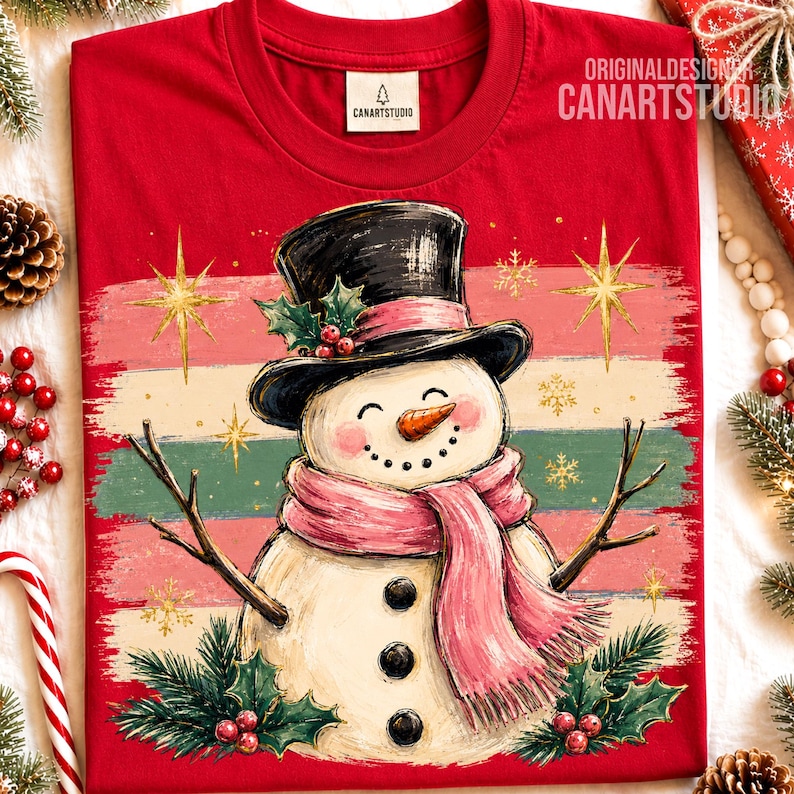 Może przedstawiać: Czerwony t-shirt z grafiką bałwana w czarnym cylindrze i r&oacute;żowym szaliku. Bałwan ma nos z marchewki, gałązkowe ramiona i trzy czarne guziki. Tło zawiera poziome paski, złote gwiazdki oraz gałązki ostrokrzewu z czerwonymi owocami.