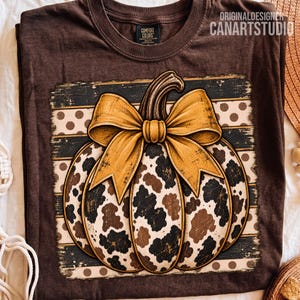 Puede incluir: Una camiseta de color marrón oscuro con un gráfico de una calabaza con estampado de vaca y un gran lazo dorado. El fondo del gráfico consiste en rayas horizontales y lunares. El texto ORIGINALDESIGNER CANARTSTUDIO es visible.