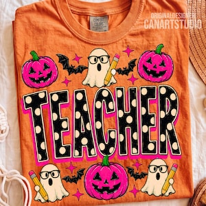 May include: An orange t-shirt featuring a graphic print with the word TEACHER in large black letters with white polka dots. The design includes three white ghosts wearing glasses and holding yellow pencils, three pink jack-o'-lanterns, and bats.