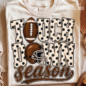 May include: A cream-coloured t-shirt featuring the text TOUCH DOWN in large block letters with a black polka dot pattern. A brown football replaces the letter O, and a brown football helmet is positioned below. The word season is in brown script.