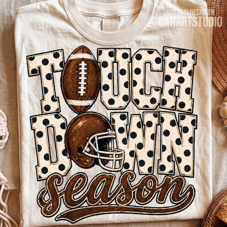 May include: A cream-coloured t-shirt featuring the text TOUCH DOWN in large block letters with a black polka dot pattern. A brown football replaces the letter O, and a brown football helmet is positioned below. The word season is in brown script.