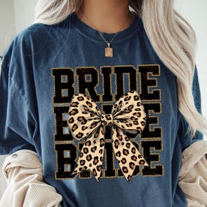 May include: A navy blue t-shirt with the text "Bride Tribe" in black with gold glitter accents. A large leopard print bow is centred on the shirt.