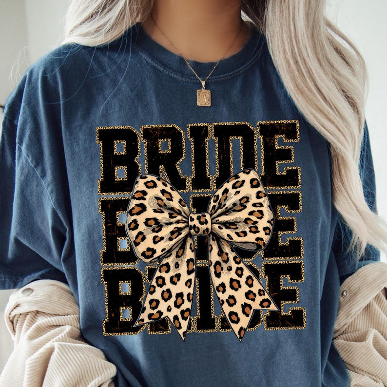 May include: A navy blue t-shirt with the text "Bride Tribe" in black with gold glitter accents. A large leopard print bow is centred on the shirt.