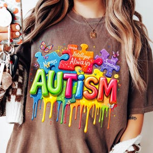 May include: A brown t-shirt with a colorful graphic of puzzle pieces and the word "AUTISM" in dripping paint. The puzzle pieces have text that reads "Unique", "Intelligent & Always", "Totally Sometimes" and "Happens".