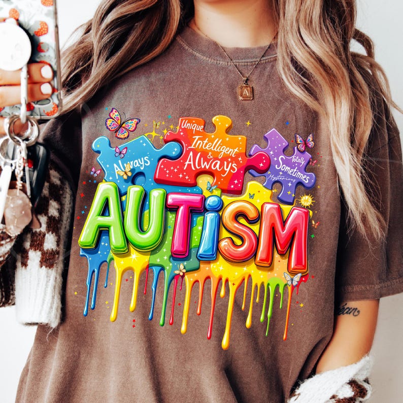 May include: A brown t-shirt with a colorful graphic of puzzle pieces and the word "AUTISM" in dripping paint. The puzzle pieces have text that reads "Unique", "Intelligent & Always", "Totally Sometimes" and "Happens".
