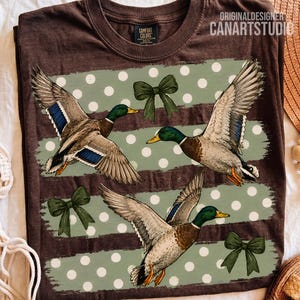 May include: A dark brown t-shirt featuring a graphic print of three flying mallard ducks with blue wing patches. The design includes horizontal sage green stripes with white polka dots and three dark green bows. The neck label reads Comfort Colors.