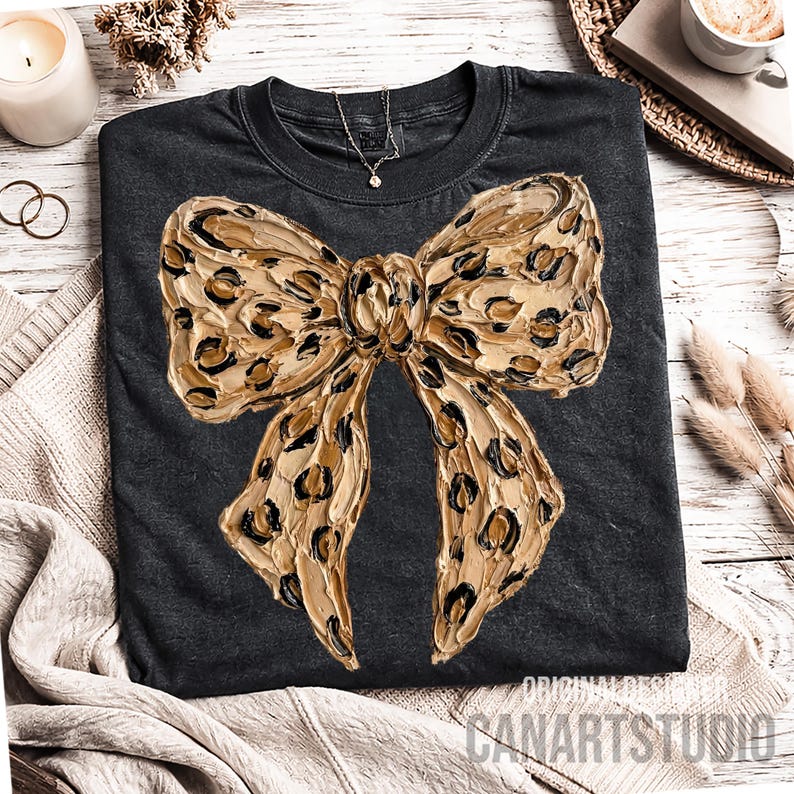 May include: Dark gray t-shirt featuring a large, textured bow design in shades of tan and black, resembling a leopard print. The bow is the focal point, with a detailed, artistic rendering. The shirt has a crew neck and short sleeves.