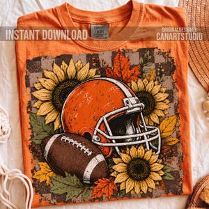 May include: An orange t-shirt featuring a graphic print of a football helmet and a football surrounded by sunflowers and autumn leaves. The design is set against a checkered background. The shirt has a Comfort Colors label at the inner neckline.