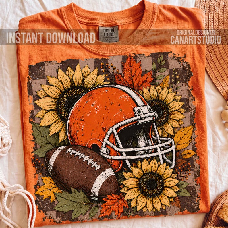May include: An orange t-shirt featuring a graphic print of a football helmet and a football surrounded by sunflowers and autumn leaves. The design is set against a checkered background. The shirt has a Comfort Colors label at the inner neckline.