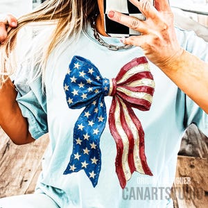 May include: Pale blue t-shirt with a large bow design in the colours of the American flag. The bow has blue sections with white stars and red and white striped sections. The text "ORIGINAL DESIGNER CANARTSTUDIO" is printed on the bottom right.