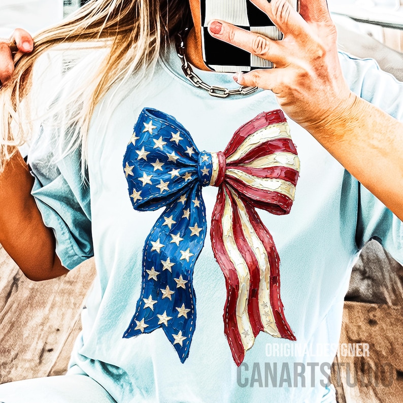 May include: Pale blue t-shirt with a large bow design in the colours of the American flag. The bow has blue sections with white stars and red and white striped sections. The text "ORIGINAL DESIGNER CANARTSTUDIO" is printed on the bottom right.