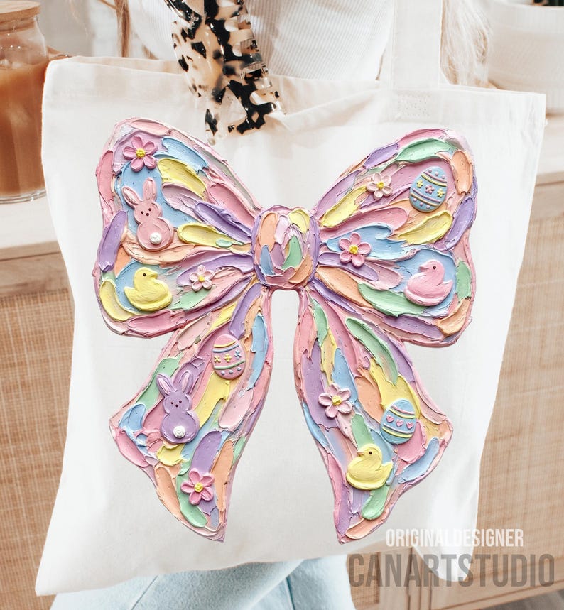 May include: White tote bag featuring a large, textured bow design in pastel colors. The bow is decorated with Easter-themed elements like eggs, flowers, bunnies, and chicks. The words "ORIGINAL DESIGNER" and "CANARTSTUDIO" are printed on the bottom right.