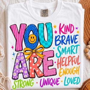 May include: A white t-shirt featuring the text YOU ARE in large, colorful, patterned letters with a yellow bow. To the right and bottom, the words KIND, BRAVE, SMART, HELPFUL, ENOUGH, STRONG, UNIQUE, and LOVED are printed in various bright colors.