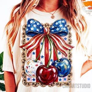 May include: Cream-colored t-shirt featuring a patriotic design. The graphic includes a blue bow with white stars, red and white stripes, and three cherries in red, white, and blue. The design is framed with a leopard print border.