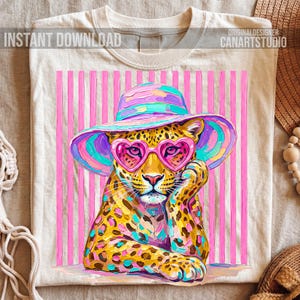 May include: Cream-colored t-shirt featuring a colorful illustration of a leopard wearing heart-shaped sunglasses and a wide-brimmed hat. The leopard is set against a backdrop of pink vertical stripes. Text on the shirt reads "INSTANT DOWNLOAD" and "ORIGINALDESIGNER CANARTSTUDIO."