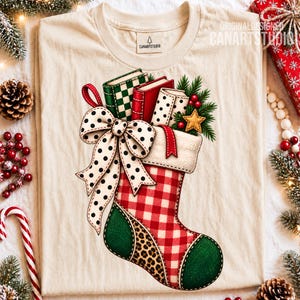 May include: A cream-colored t-shirt featuring a graphic of a Christmas stocking with a red and white gingham pattern, a green toe and heel, and a leopard print section. The stocking is filled with books, pine branches, and a gold star.