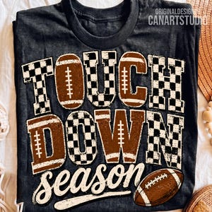 May include: A dark gray t-shirt featuring the text TOUCH DOWN season in a distressed font. The letters are filled with a brown football texture and a black and white checkered pattern. A brown football graphic is positioned at the bottom right.