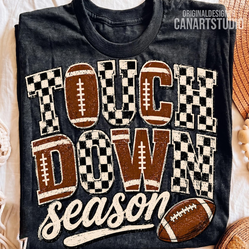 May include: A dark gray t-shirt featuring the text TOUCH DOWN season in a distressed font. The letters are filled with a brown football texture and a black and white checkered pattern. A brown football graphic is positioned at the bottom right.
