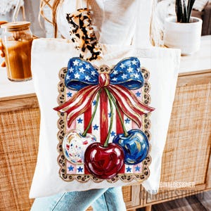 May include: White canvas tote bag featuring a patriotic design. The design includes a blue bow with white stars, red and white striped ribbons, and three cherries. The cherries are painted in red, blue, and white with star accents. The bag has a leopard print handle.