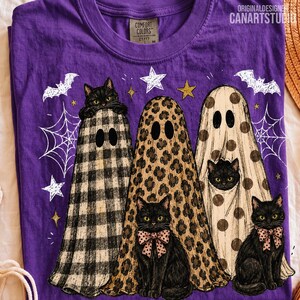 May include: A purple t-shirt featuring a graphic of three black cats wearing ghost costumes with different patterns including black and white buffalo plaid, leopard print, and brown polka dots, surrounded by white spiderwebs, stars, and bats.